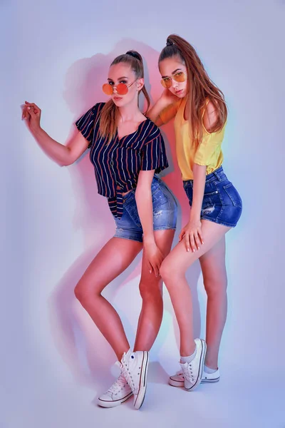Two female teenagers in sunglasses and summer clothes posing in studio on white background — Stock Photo, Image