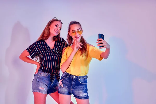 Dos adolescentes en ropa de verano de pie en el estudio y haciendo selfie sobre fondo blanco. Luz mixta — Foto de Stock