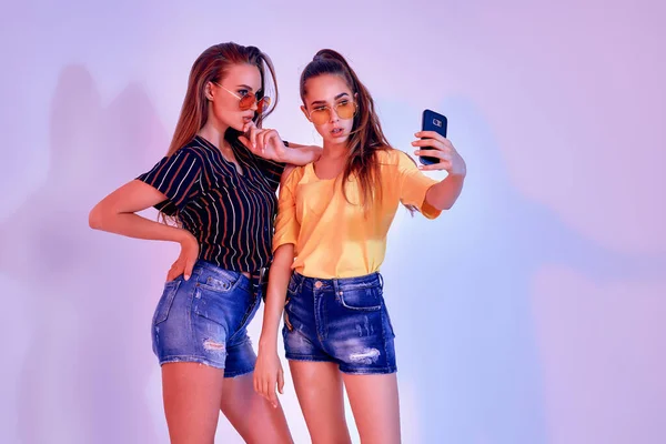 Dvě dospívající dívky v letním oblečení stojící ve studiu a dělat selfie na bílém pozadí. Smíšené světlo — Stock fotografie