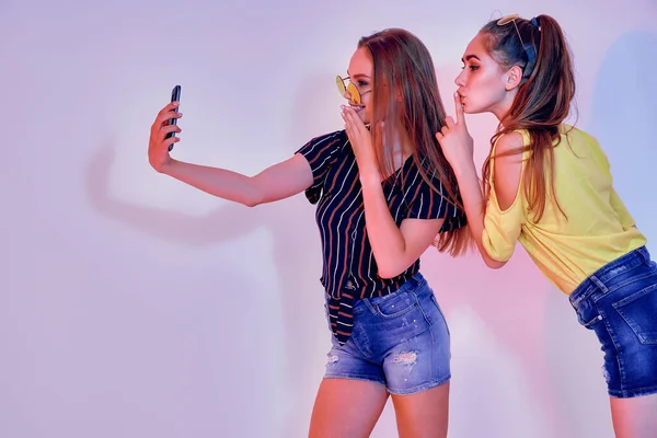 Dos adolescentes en ropa de verano de pie en el estudio y haciendo selfie sobre fondo blanco. Luz mixta —  Fotos de Stock