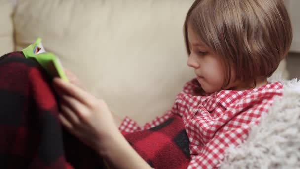 Liten flicka 9 år gammal läser en bok när hon ligger på soffan. — Stockvideo