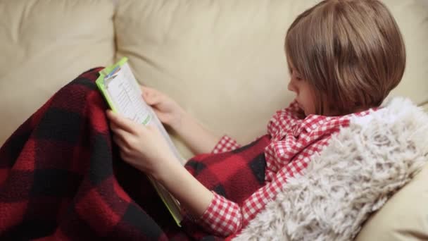 A 9 éves kislány könyvet olvas, miközben a kanapén fekszik.. — Stock videók