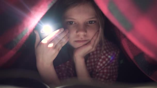 A cute little girl, hiding under the bed sheet in the bedroom, is happily reading a book with the help of the light. — Stock Video