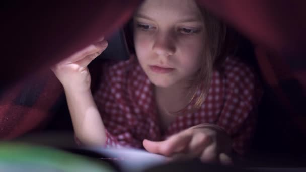 Una niña linda, escondida debajo de la sábana en el dormitorio, está felizmente leyendo un libro con la ayuda de la luz. — Vídeos de Stock