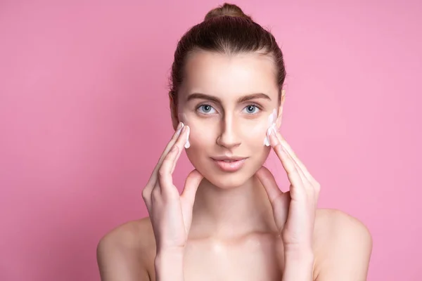 Mooie jonge vrouw aanbrengen crème op haar gezicht tegen kleur achtergrond — Stockfoto