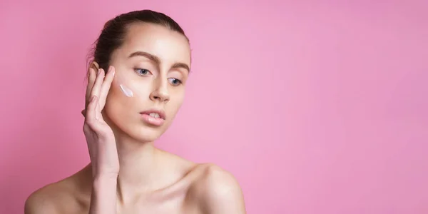 Bella giovane donna applicando crema sul suo viso contro lo sfondo di colore — Foto Stock