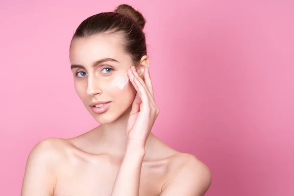 Beautiful young woman applying cream on her face against color background — Stock Photo, Image