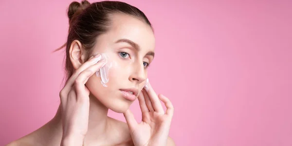 Bella giovane donna applicando crema sul suo viso contro lo sfondo di colore — Foto Stock