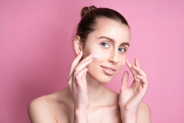 Mooie jonge vrouw aanbrengen crème op haar gezicht tegen kleur achtergrond — Stockfoto