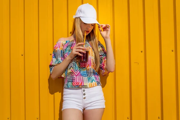 Bastante joven chica estilo hipster divertirse al aire libre en gorra blanca y comer helado sobre fondo amarillo estilo urbano —  Fotos de Stock