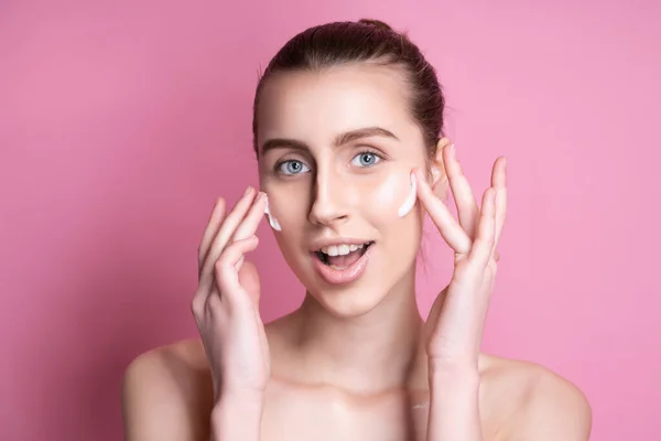 Gyönyörű fiatal nő alkalmazása krém az arcára ellen színes háttér — Stock Fotó