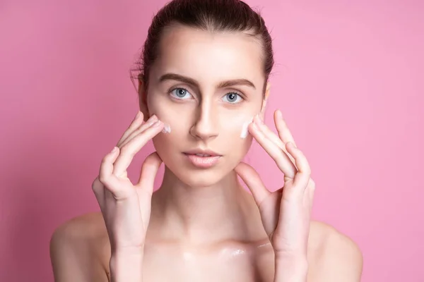 Mooie jonge vrouw aanbrengen crème op haar gezicht tegen kleur achtergrond — Stockfoto