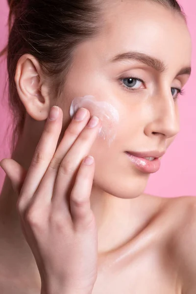 Mooie jonge vrouw aanbrengen crème op haar gezicht tegen kleur achtergrond — Stockfoto