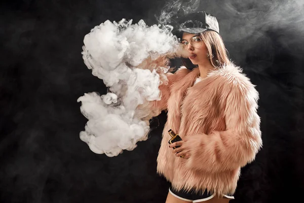 Sexy trendy rap vrouw in roze bontjas en pet rook elektronische sigaret. Dark Studio achtergrond schieten — Stockfoto