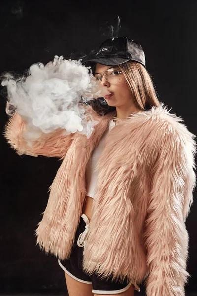 Sexy mulher rap na moda em casaco de pele rosa e cigarro eletrônico tampa fumaça. Estúdio escuro tiro fundo — Fotografia de Stock