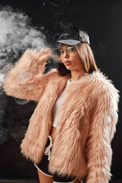 Сексуальная модная рэп-женщина в розовом шубе и кепке курит электронную сигарету. Фотосессия Dark Studio — стоковое фото