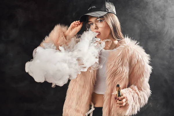 Sexy trendy rap woman in pink fur coat and cap smoke electronic cigarette. Dark Studio background shooting — Stock Photo, Image