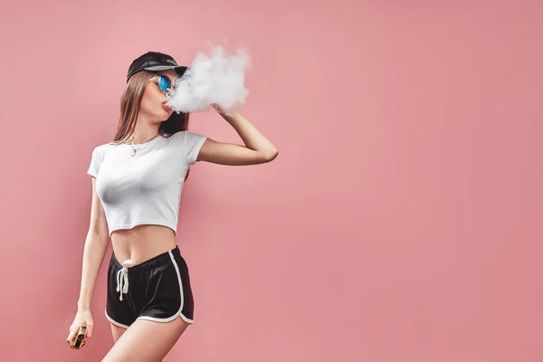 Mujer joven seductora glamour de pie y vapeo sobre fondo rosa — Foto de Stock