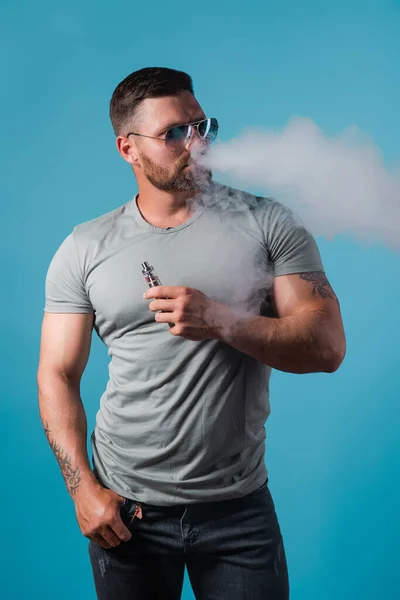 タバコに代わるものとして蒸気タバコを吸うサングラスでひげを生やした残忍な男性。スタジオ撮影青の背景. — ストック写真