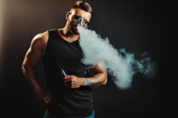 Varón barbudo con gafas de sol fumando un cigarrillo de vapor como alternativa al tabaco. Estudio sobre fondo oscuro. — Foto de Stock