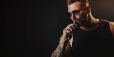 Tütüne alternatif olarak sigara içen güneş gözlüklü vahşi erkek. Stüdyo çekimi koyu arkaplanda.