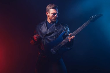 Deri ceketli ve güneş gözlüklü vahşi sakallı heavy metal müzisyeni elektro gitar çalıyor. Karanlık arka planda bir stüdyoda dumanla çekilmiş.