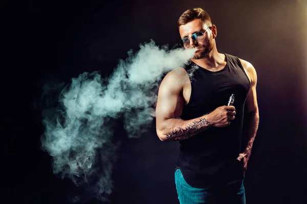 Bearded brutal male in sunglasses smoking a vapor cigarette as an alternative to tobacco. Studio shot on dark background. — Stock Photo, Image