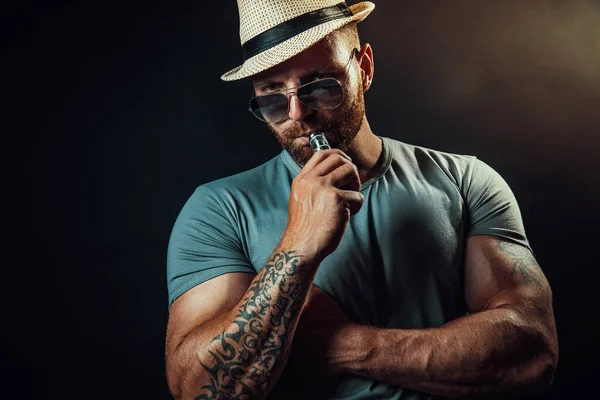 Brutalny mężczyzna w kapeluszu i okularach przeciwsłonecznych palący papierosa jako alternatywę dla tytoniu. Studio nakręcone na ciemnym tle. — Zdjęcie stockowe