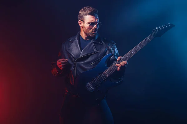 Brutal bearded Heavy metal musician in leather jacket and sunglasses is playing electrical guitar. Shot in a studio on dark background with smoke — Stock Photo, Image