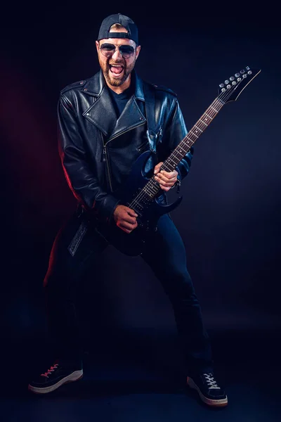 Teljes méretű fotó brutális szakállas Heavy metal zenész bőrdzsekiben és napszemüvegben játszik elektromos gitáron nagyon érzelmileg. Lelőtték egy stúdióban a sötét háttér — Stock Fotó