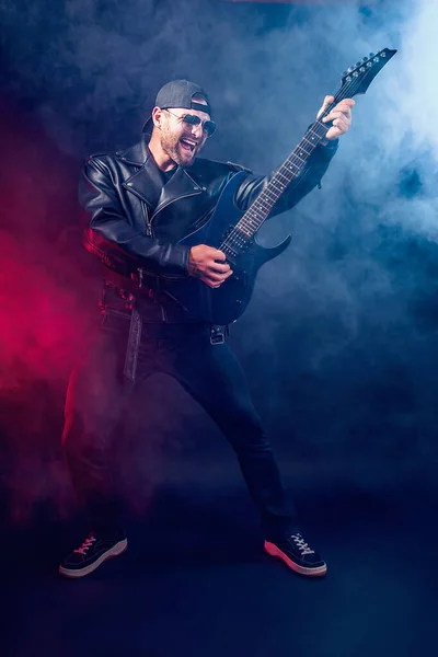 Full size photo of brutal bearded Heavy metal musician in leather jacket and sunglasses is playing electrical guitar very emotionally. Shot in a studio on dark background with smoke — Stock Photo, Image