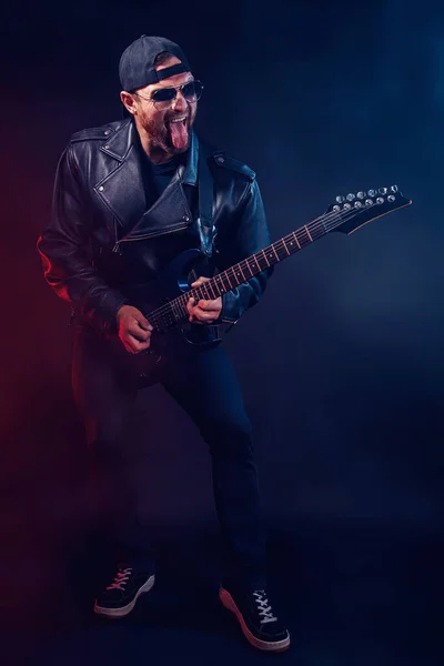 Pełnowymiarowe zdjęcie brutalnego brodatego muzyka heavy metalowego w skórzanej kurtce i okularach przeciwsłonecznych gra na gitarze elektrycznej bardzo emocjonalnie. Kręcony w studiu na ciemnym tle. — Zdjęcie stockowe