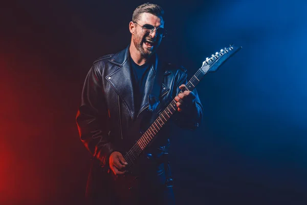 Brutal bearded Heavy metal musician in leather jacket and sunglasses is playing electrical guitar. Shot in a studio on dark background with smoke — Stock Photo, Image