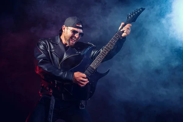 Brutal bearded Heavy metal musician in leather jacket and sunglasses is playing electrical guitar. Shot in a studio on dark background with smoke — Stock Photo, Image