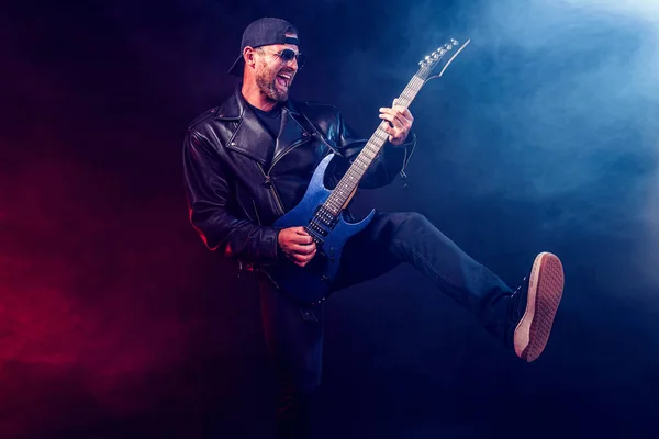 Brutal bearded Heavy metal musician in leather jacket and sunglasses is playing electrical guitar. Shot in a studio on dark background with smoke — Stock Photo, Image