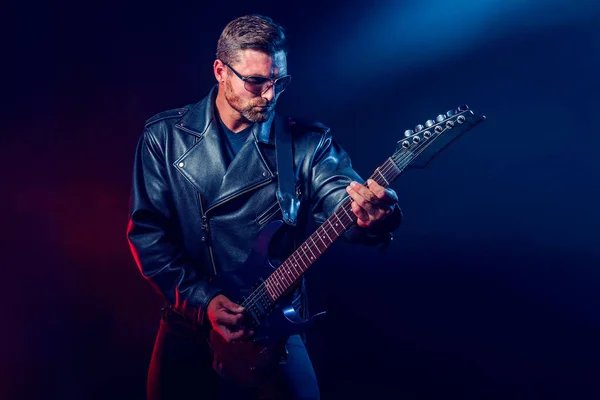 Brutal bearded Heavy metal musician in leather jacket, cap and sunglasses is playing electrical guitar. Shot in a studio on dark background — Stock Photo, Image