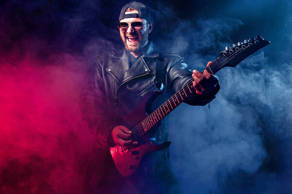 Жестокий бородатый металлический музыкант в кожаной куртке и солнечных очках играет на электрогитаре. Съемка в студии на темном фоне с дымом