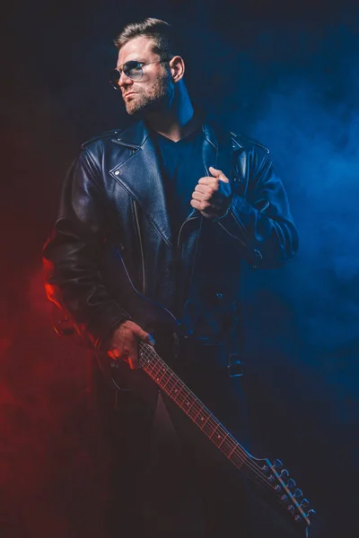 Brutal bearded Heavy metal musician in leather jacket, cap and sunglasses is playing electrical guitar. Shot in a studio on dark background — Stock Photo, Image