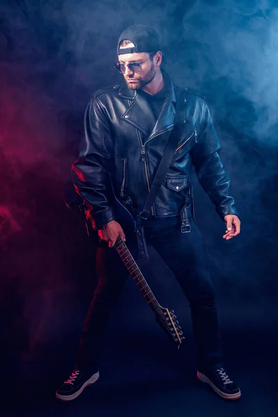 Full size photo of brutal bearded Heavy metal musician in leather jacket and sunglasses with electrical guitar. Shot in a studio on dark background with smoke