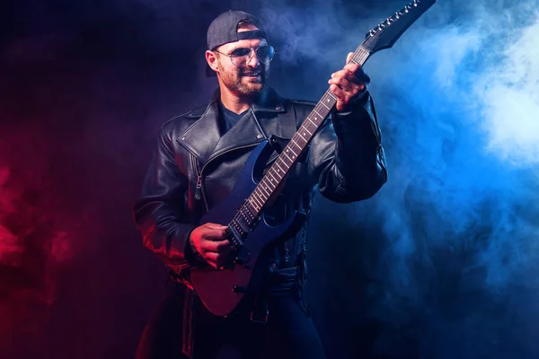 Brutal bearded Heavy metal musician in leather jacket and sunglasses is playing electrical guitar. Shot in a studio on dark background with smoke — Stock Photo, Image