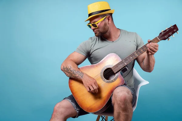 Handsome bearded guitarist in yellow hat and sunglasses sitting on chair and singing while playing on acoustic guitar isolated on blue background — Stock Photo, Image