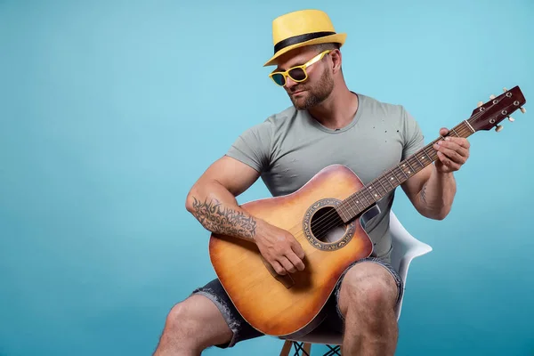 Handsome bearded guitarist in yellow hat and sunglasses sitting on chair and singing while playing on acoustic guitar isolated on blue background — Stock Photo, Image