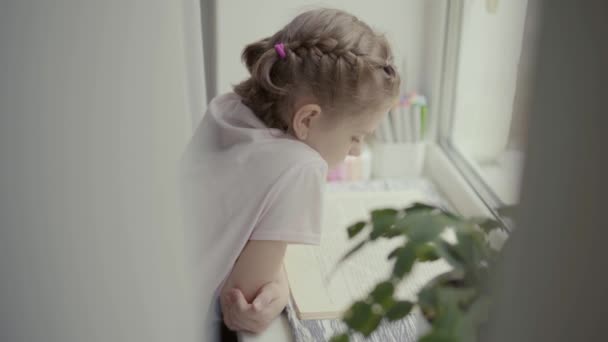 Vacker liten flicka läser en bok som sitter nära ett fönster, regn utanför — Stockvideo