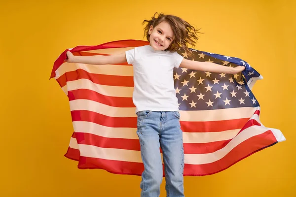 Glad barn, söt flicka med amerikansk flagga på gul studio bakgrund. USA firar 4 juli, självständighetsdagen. — Stockfoto