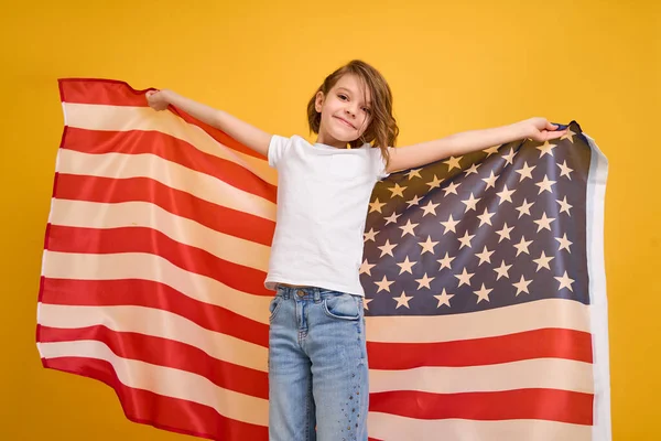 Glad barn, söt flicka med amerikansk flagga på gul studio bakgrund. USA firar 4 juli, självständighetsdagen. — Stockfoto