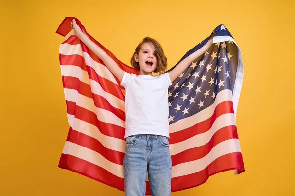 Glad barn, söt flicka med amerikansk flagga på gul studio bakgrund. USA firar 4 juli, självständighetsdagen. — Stockfoto