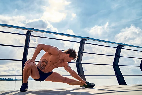 Νεαρός μυώδης άνδρας με τατουάζ κάνει ασκήσεις, που τεντώνεται νωρίς το πρωί στην προβλήτα δίπλα στη θάλασσα — Φωτογραφία Αρχείου