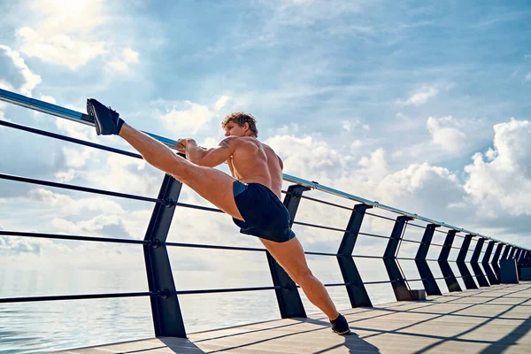 Jeune homme tatoué musclé faisant des exercices, s'étirant tôt le matin sur la jetée près de la mer — Photo