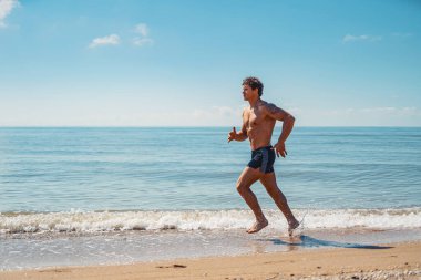 Genç kaslı adam, plajda çıplak bir gövde ile koşuyor.
