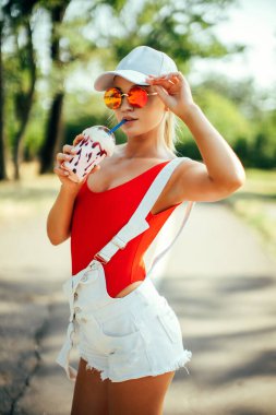 Yuvarlak güneş gözlüğü takan seksi moda kızı portresi, kırmızı mayo, beyaz beyzbol şapkası, açık havada milkshake 'li şort.. 